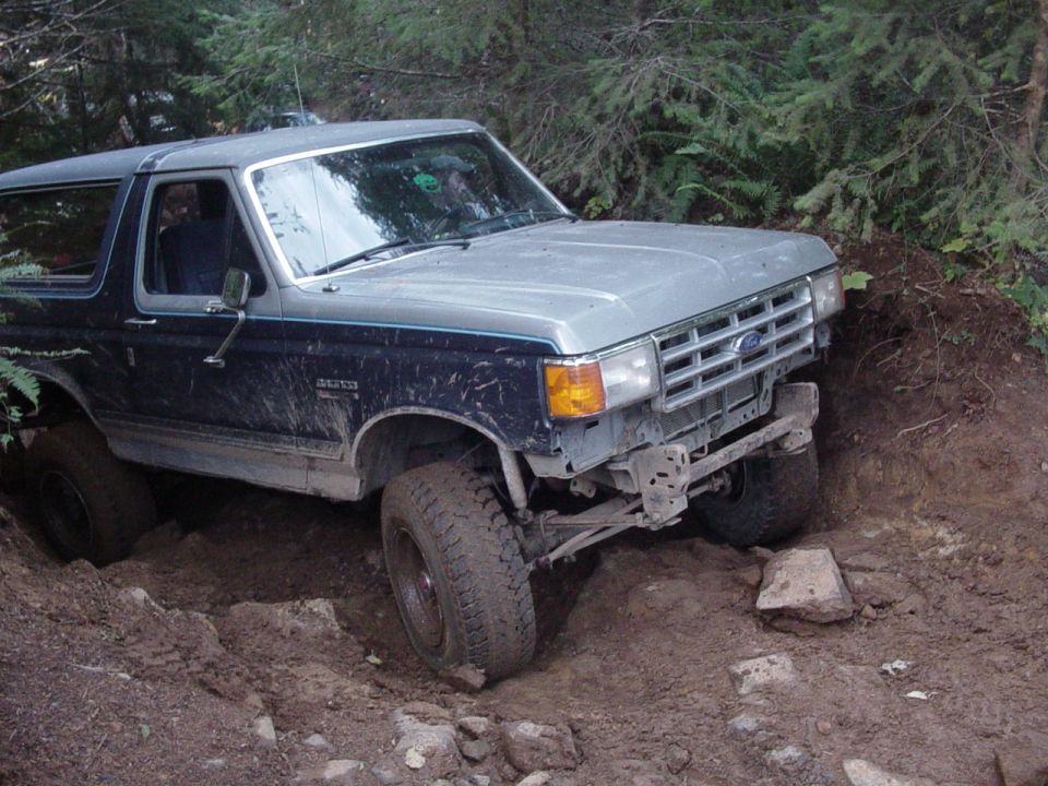 Two-tone blue and silver Ford Bronco flexing in a muddy rutted trail with front bumper removed