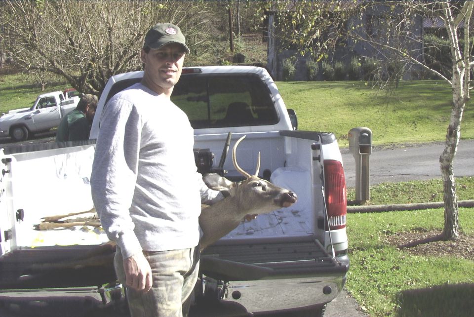 Man with deer in pickup truck bed. Another truck is visible in the background