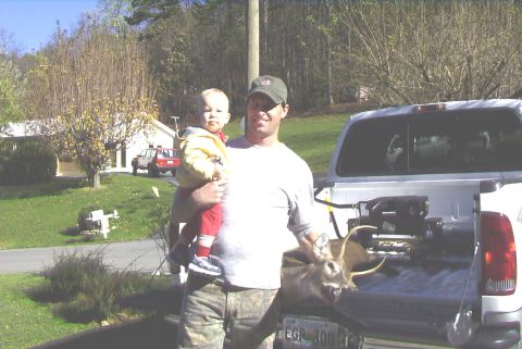 1995 Ford Bronco - A man holds a baby while standing next to a truck bed containing a deer, with two red SUVs visible in the background.