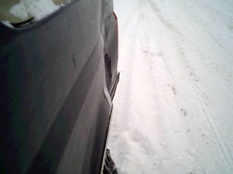 1987 Ford Bronco - A dark SUV's rear quarter panel is visible in a snowy environment, with tire tracks in the snow.