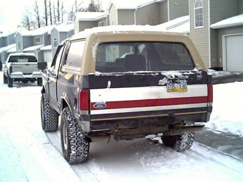 1987 Ford Bronco - A 1987 Ford Bronco is parked in the snow, showing its rear end and a white pickup truck in the background.