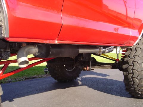 1978 Ford Bronco - Low ground-level view under the rocker of a lifted red truck, showing a side-exit exhaust with a large muffler and turned-down tailpipe rou…