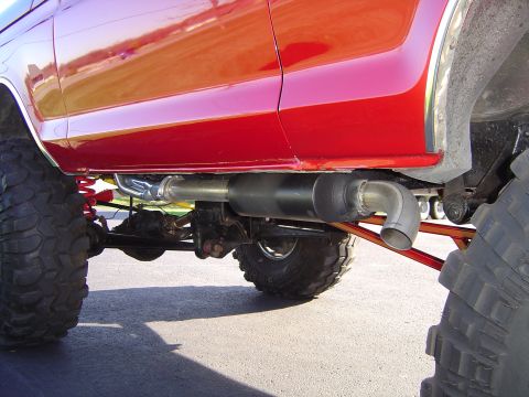 1978 Ford Bronco - Ground-level view beneath the rocker of a lifted red truck showing a stainless exhaust with a large polished muffler and side-exit turndown…