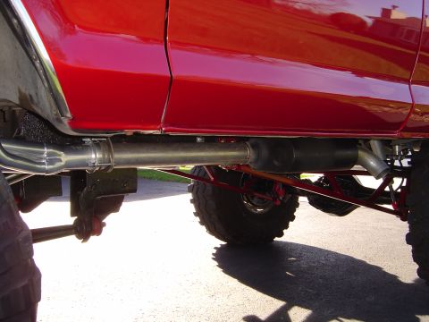 1978 Ford Bronco - Low sunlit view along the rocker of a lifted red truck, showing a full-length side exhaust running from a header collector through a black…