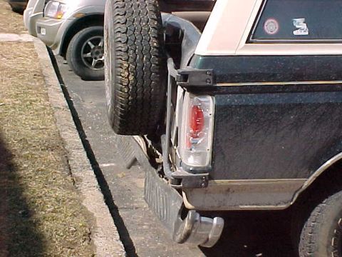 1993 Ford Bronco - Dark green SUV with spare tire on back, parked next to a silver SUV. Features a tow hitch and visible taillight.