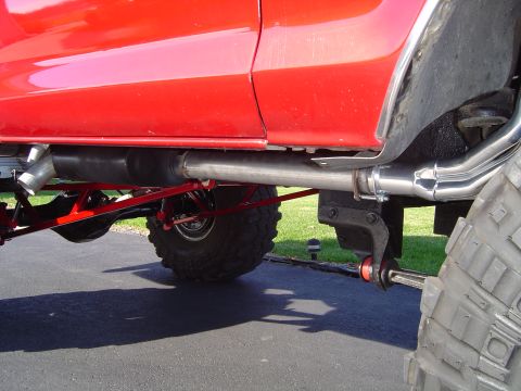 1978 Ford Bronco - Low ground-level view under the side of a lifted red truck body, showing polished stainless exhaust tubing with a muffler routed over the f…