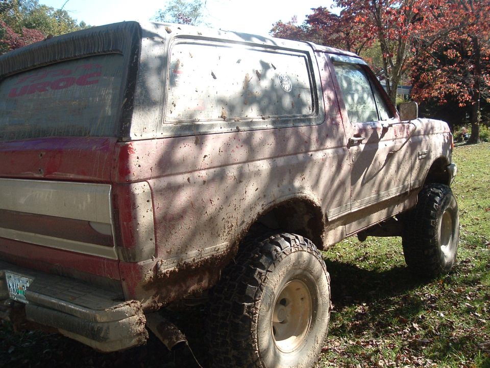 Red and white 1987 Ford Bronco covered in mud, lifted with large tires, viewed from the right rear