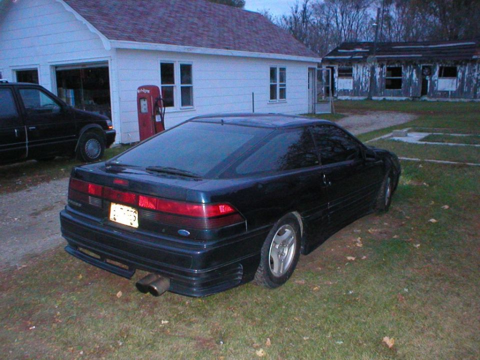 Dark green coupe rear 3/4 view parked on grass near a white house and vintage gas pump