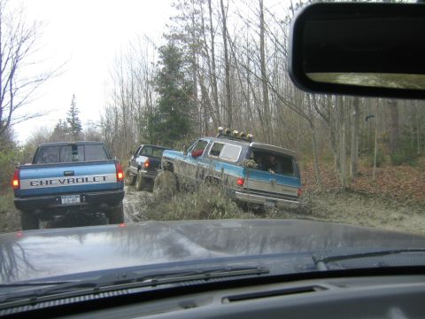 1994 Ford Bronco - Several trucks navigate a muddy trail through a wooded area, with one SUV deeply mired in the muck.