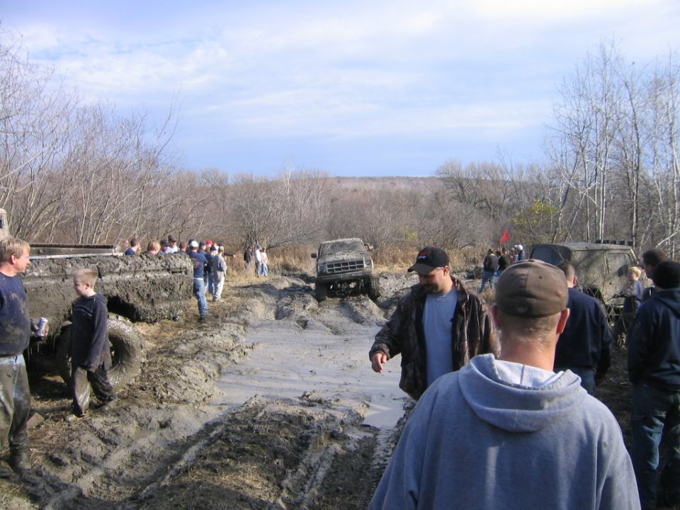 Muddy off-road vehicles stuck in a bog with spectators watching. Some vehicles are partially submerged in mud