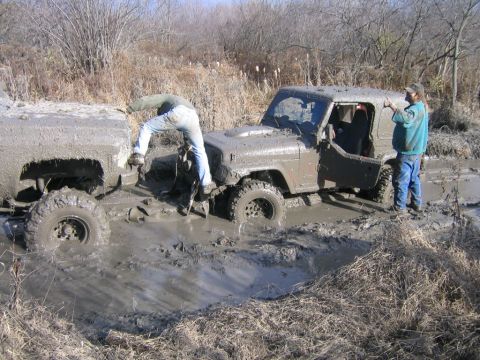 1994 Ford Bronco - Two off-road vehicles are deeply stuck in mud, with individuals attempting to free them from the swampy terrain.
