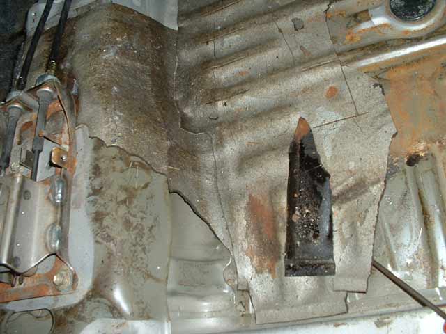 Stripped car interior floor pan with torn insulation, surface rust, and a rusty bracket on the tunnel