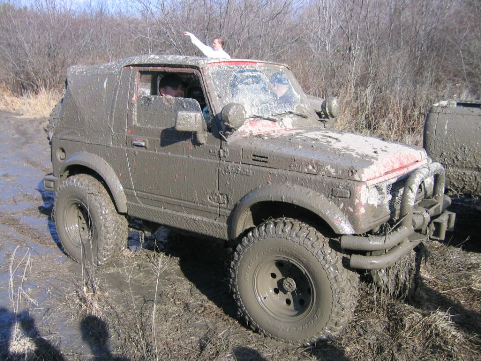 Mud-covered Suzuki Samurai off-roading, with a person on the roof pointing