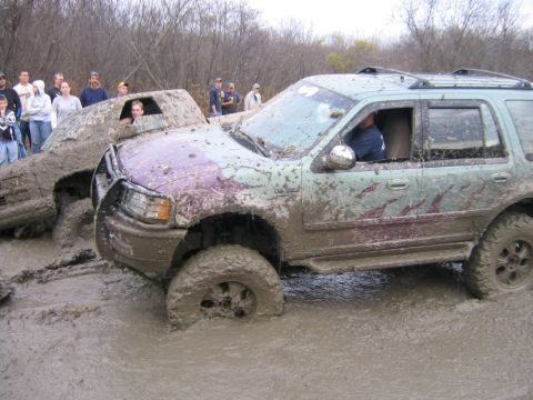 1994 Ford Bronco - Off-road vehicles are deeply mired in thick mud, with onlookers gathered on the bank.