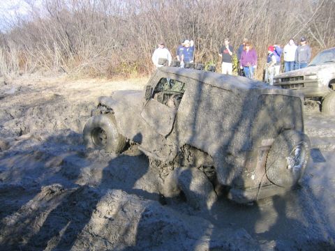 1994 Ford Bronco - An off-road vehicle is deeply stuck in mud during a poker run event, with spectators observing the scene.