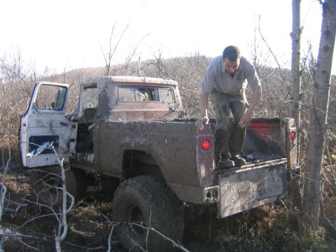 1994 Ford Bronco - A man climbs into the bed of a heavily mud-covered pickup truck stuck in a wooded area.