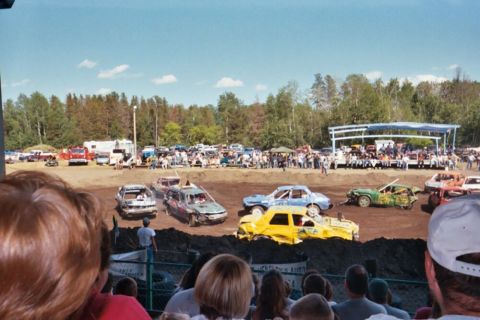 1996 Ford Bronco - Several beat-up cars are in a dirt arena, possibly during a demolition derby event, with a crowd watching.