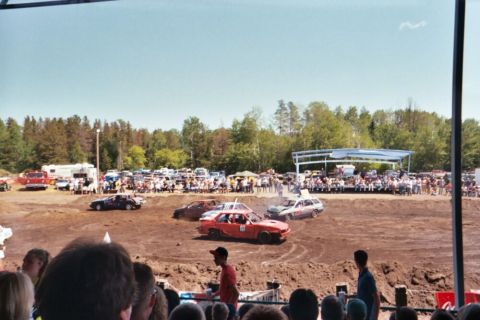 1996 Ford Bronco - A demolition derby is underway with several cars in a dirt arena, and a crowd watching the event.