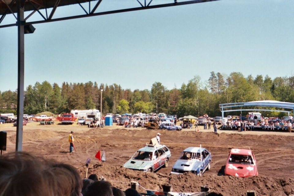 Demolition derby cars stuck in mud under a bright sky, with spectators in the background