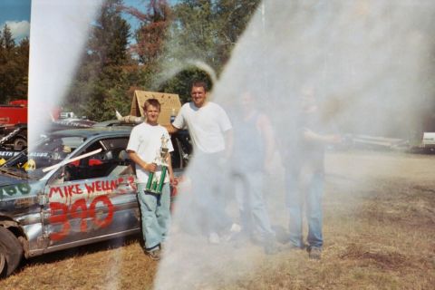 1996 Ford Bronco - Two men pose with a trophy next to a heavily damaged car at a demolition derby event.