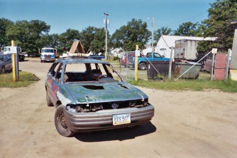 1996 Ford Bronco - This heavily modified sedan, likely prepared for demolition derby, shows significant damage to its body and roof, with unusual decorations…