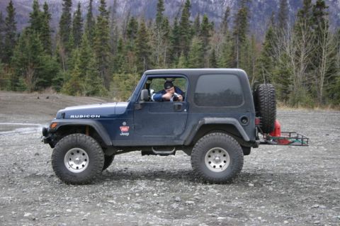 2003 Jeep TJ - A blue Jeep Wrangler is parked on a gravel surface with trees and mountains in the background.