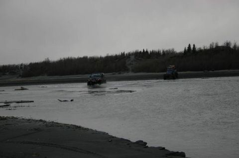 2003 Jeep TJ - Two vehicles traverse a shallow body of water with a treeline in the distance under a cloudy sky.