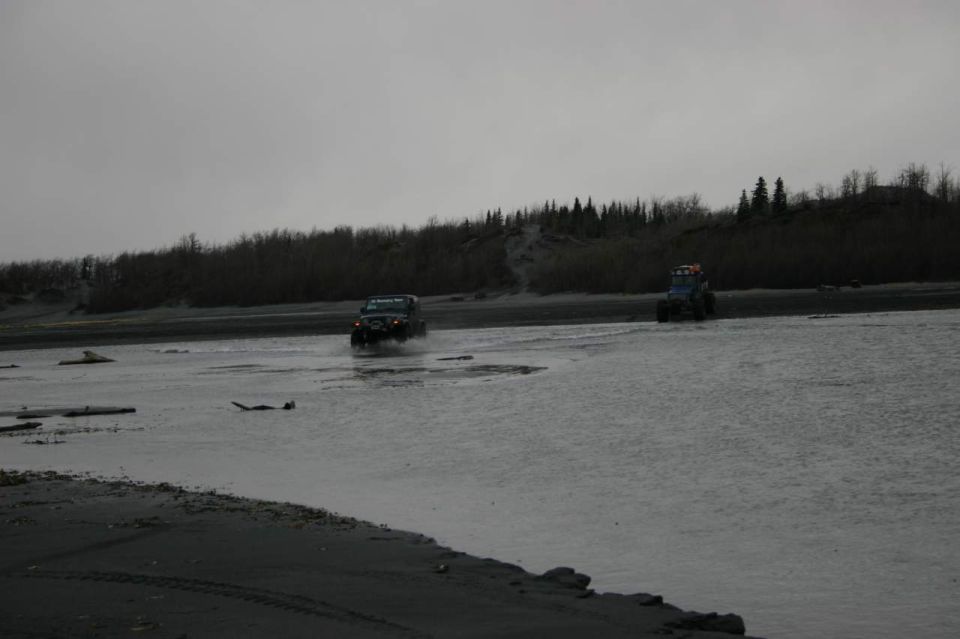 Dark colored Jeep TJ driving through water, front view, with a blue tractor in the background