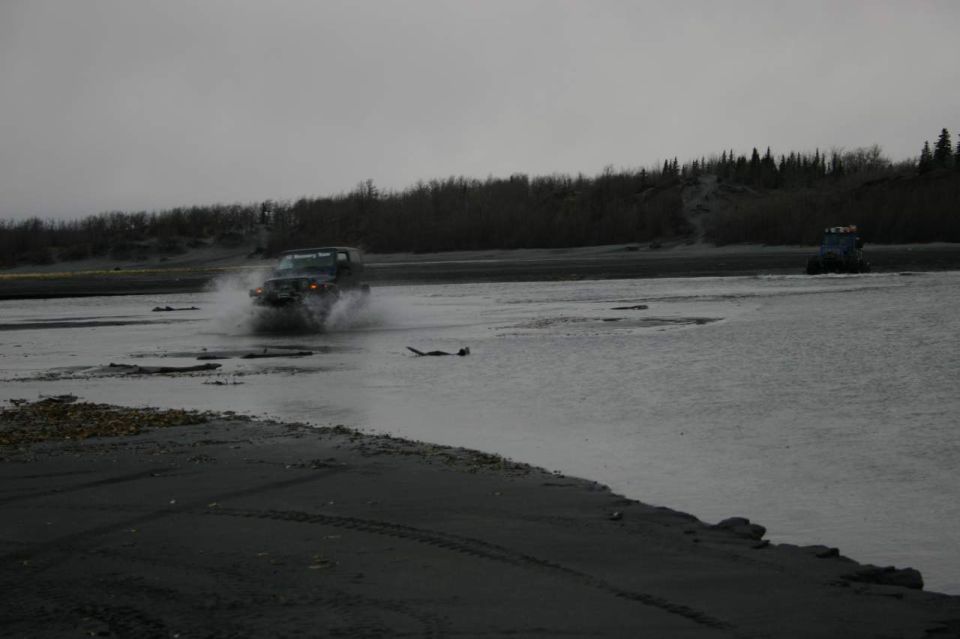 Dark Jeep Wrangler splashing through a shallow river channel on a gravel bar under overcast sky