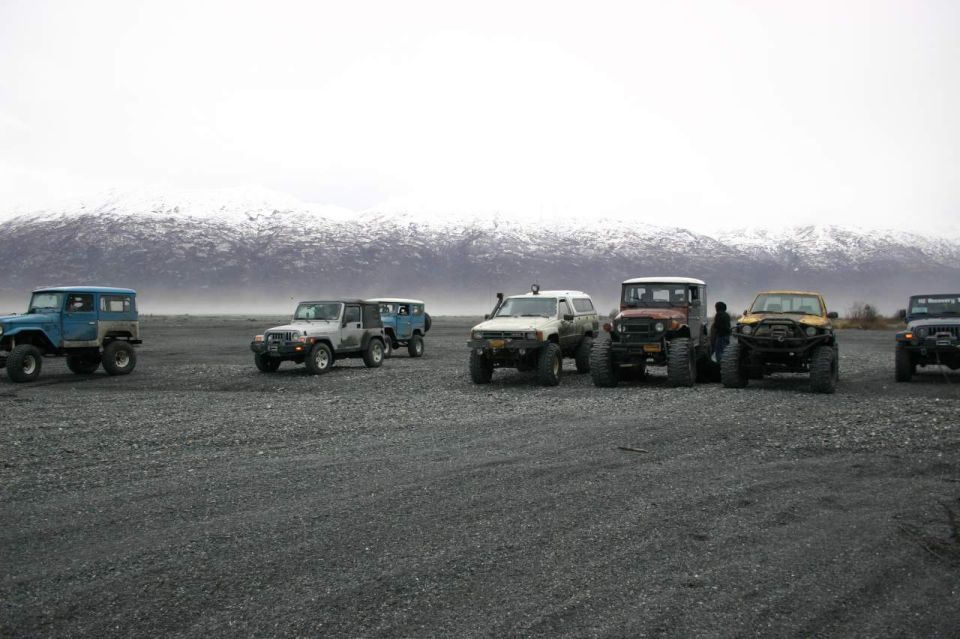Several off-road vehicles including Jeeps and older SUVs parked on a gravel plain with snow-capped mountains in the background
