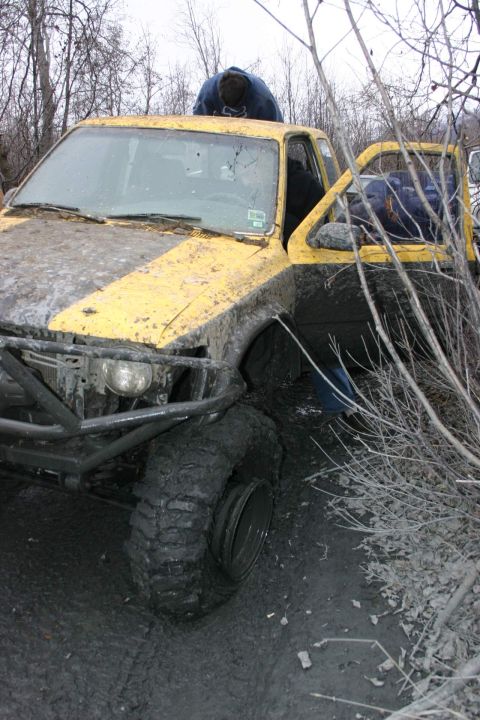 2003 Jeep TJ - A heavily mud-splattered yellow pickup truck is stuck in a muddy, wooded area.
