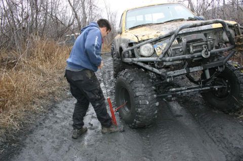 2003 Jeep TJ - A person uses a jack to lift a heavily modified off-road truck stuck in the mud.