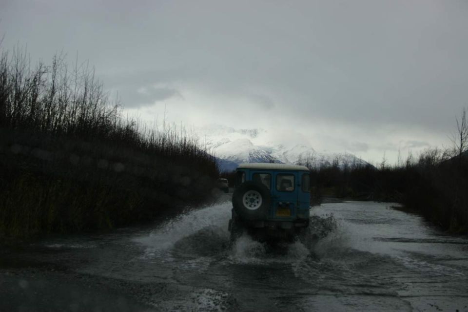 Blue off-road vehicle driving through a shallow river, kicking up water. Snow-capped mountains are in the background