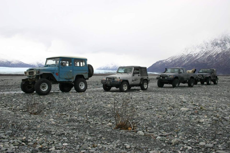 Several modified off-road vehicles parked on a rocky plain with a glacier and snow-capped mountains in the background