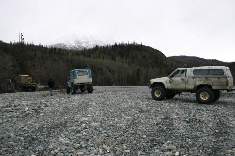 2003 Jeep TJ - A white pickup truck with a camper shell sits on a rocky surface with other vehicles and a mountain in the background.