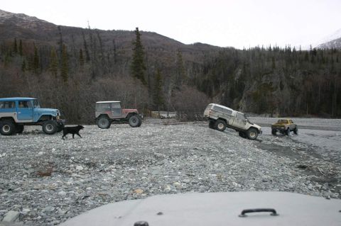 2003 Jeep TJ - Multiple off-road vehicles traverse a , rocky terrain under a cloudy sky, with dense trees and a mountain range visible.