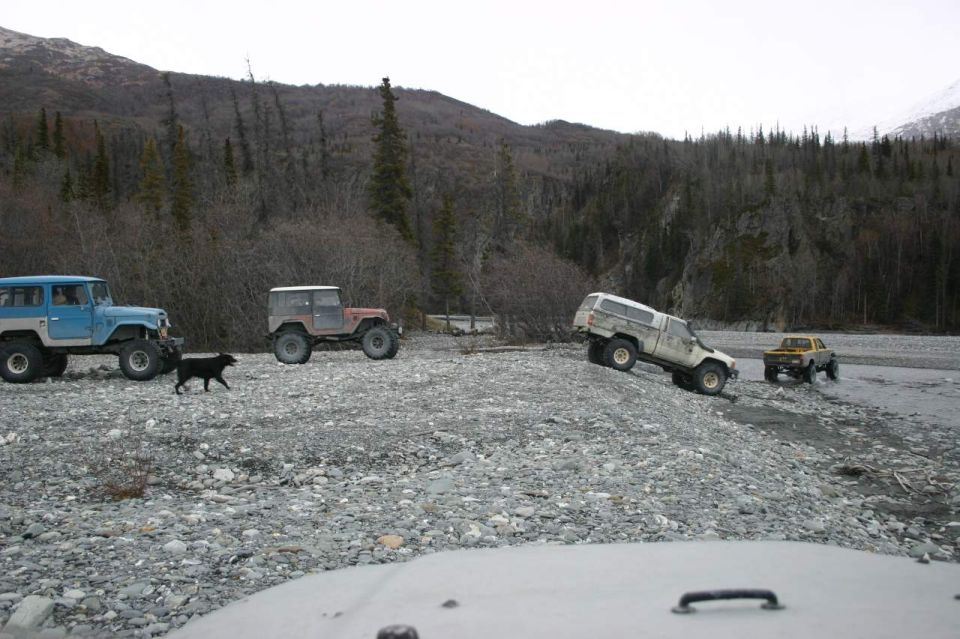 Four off-road vehicles navigate a rocky, barren landscape with trees and mountains in the background