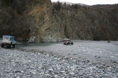 2003 Jeep TJ - Two vintage off-road vehicles navigate a wide, stony riverbed under a cloudy sky with a large rock formation behind them.