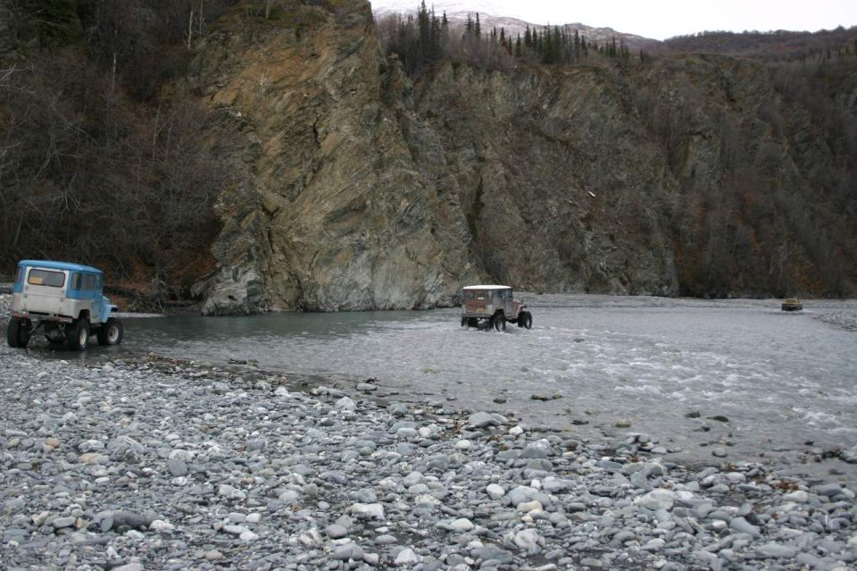 Two old 4x4s driving through a shallow, rocky river with a steep, wooded cliff face in the background