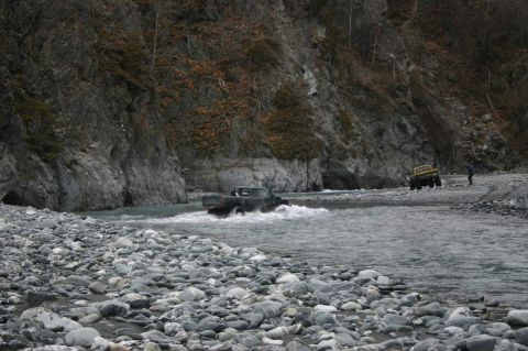 2003 Jeep TJ - Two off-road vehicles navigate a rocky riverbed, with one truck splashing through the water.