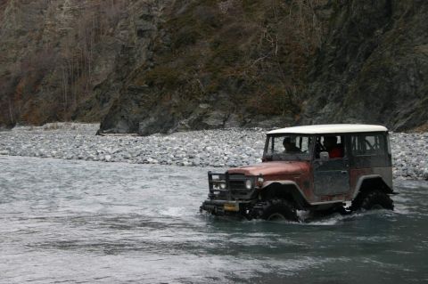 2003 Jeep TJ - This classic Toyota Land Cruiser navigates a shallow river, its tires churning water amidst a rocky landscape.