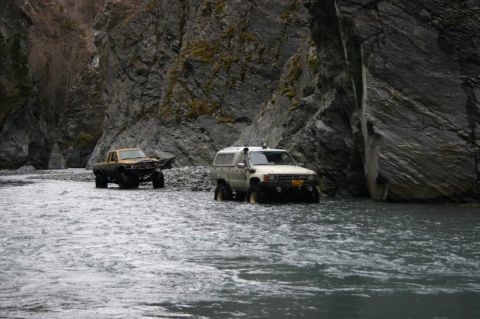 2003 Jeep TJ - Two modified trucks drive through a river in a canyon.