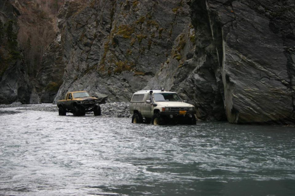 Two off-road trucks navigate a rocky riverbed within a steep canyon