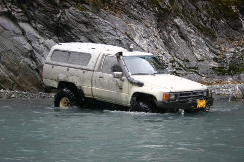 2003 Jeep TJ - This Toyota pickup truck is equipped with a snorkel and camper shell, navigating a rocky riverbed.