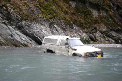 2003 Jeep TJ - A white pickup truck with a camper shell and snorkel is partially submerged in a river, with a rocky cliff face behind it.