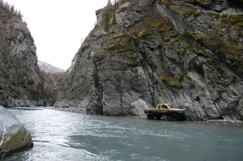 2003 Jeep TJ - A yellow pickup truck navigates a riverbed flanked by steep, rocky canyon walls.