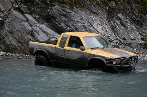 2003 Jeep TJ - A yellow pickup truck navigates a rocky riverbed, its tires submerged in the water.