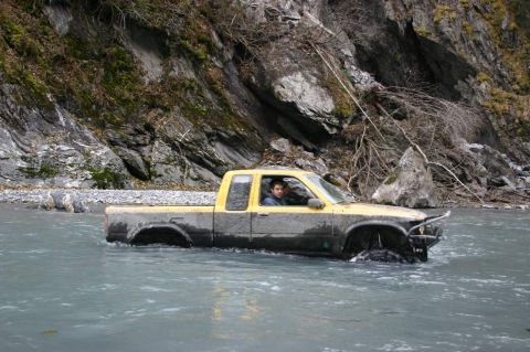2003 Jeep TJ - A yellow pickup truck navigates through a shallow river with rocky banks and dense vegetation.