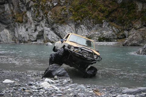 2003 Jeep TJ - A modified yellow truck attempts to cross a shallow, rocky river with steep, rocky banks.