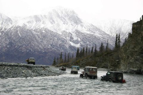 2003 Jeep TJ - Several off-road vehicles navigate a rocky riverbed beneath a , snow-covered mountain range.